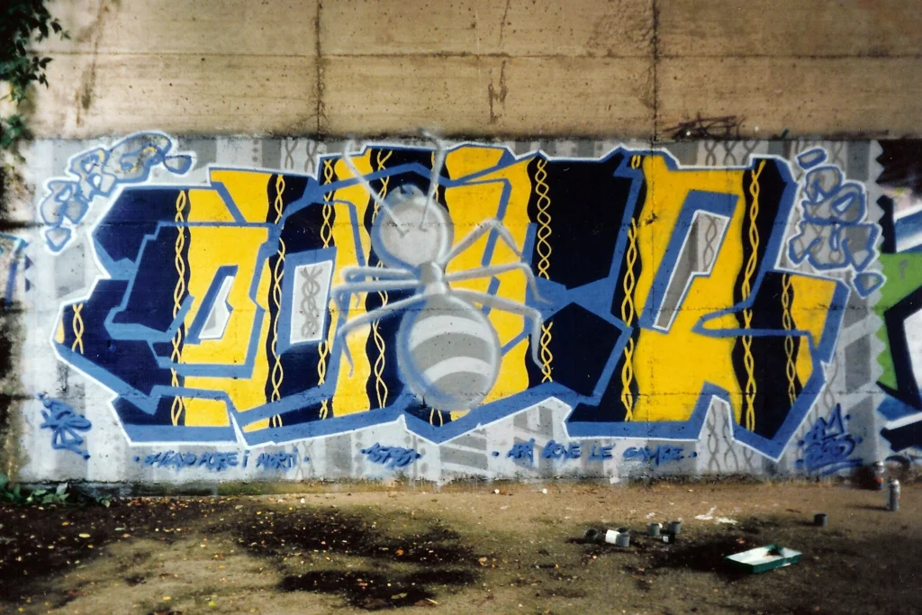 Graffiti su muro di cemento con lettere blu e gialle e una grande formica grigia dipinta al centro.