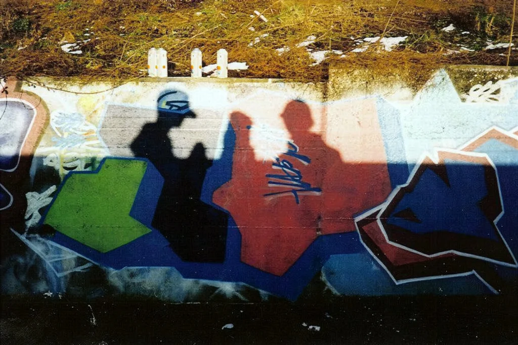 Graffiti colorato con lettere geometriche e ombre di writer proiettate sul muro.