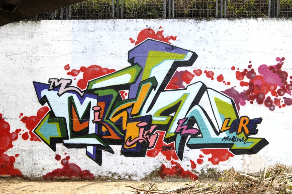 Graffiti con lettering verde, viola e arancio su muro bianco, Tradate 2006.